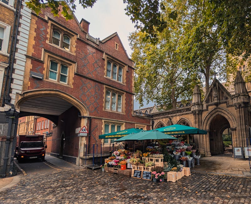 An outdoor flower stall situated under a green umbrella on a cobblestone street near a historic brick building with an arched entrance and Gothic architectural details. The stall displays a vibrant ar