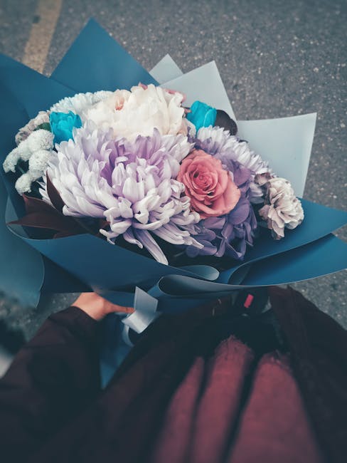 A hand holding a bouquet of fresh flowers wrapped in dark blue paper, featuring a pink rose, large light purple chrysanthemums, white flowers, and small bright blue blossoms, arranged in a compact, ro