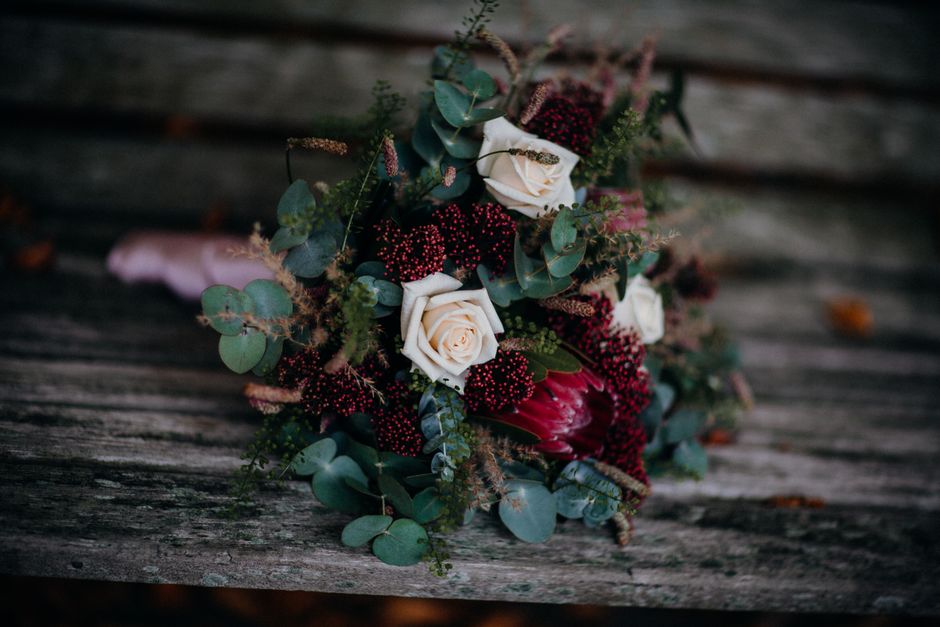 A floral arrangement featuring a bouquet with creamy white roses, deep red and burgundy blooms, and sprigs of eucalyptus with round, green leaves, all arranged in a loose, natural style. The bouquet r