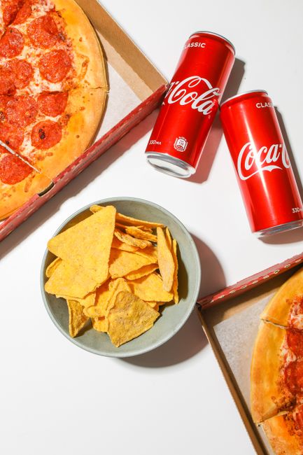 A flat lay image featuring two partially visible pizza slices in cardboard boxes, with golden-brown crusts and melted cheese toppings. Positioned centrally is a small grey bowl filled with yellow corn