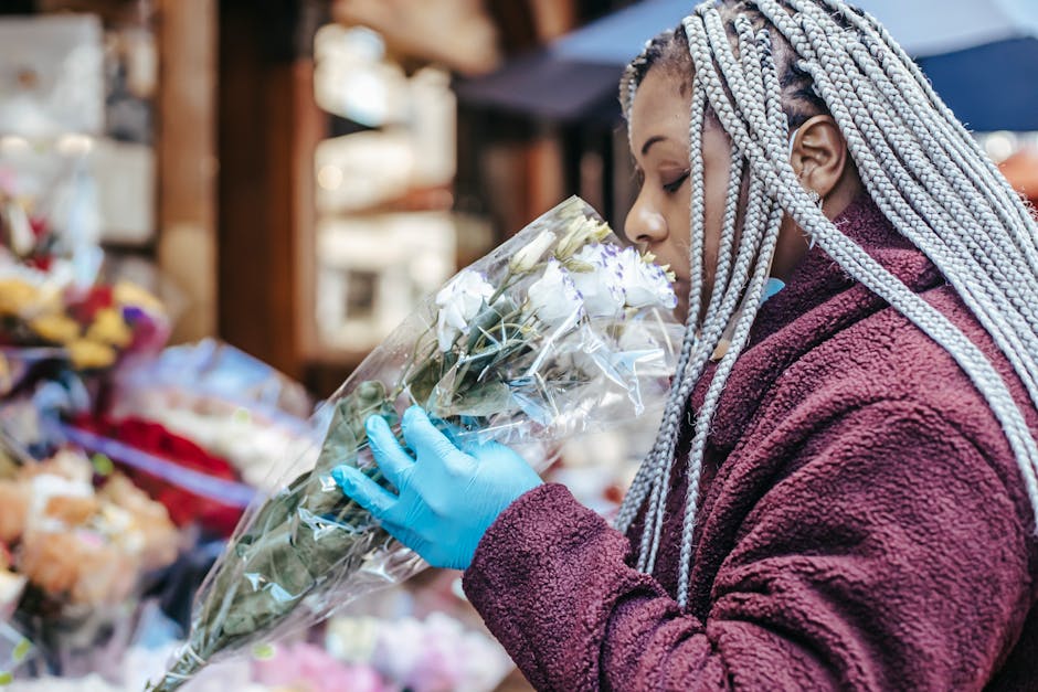 A woman with silver braided hair, wearing a maroon fleece jacket and blue gloves, closely smelling a bouquet of white roses wrapped in clear plastic at an outdoor flower market. The arrangement is hel